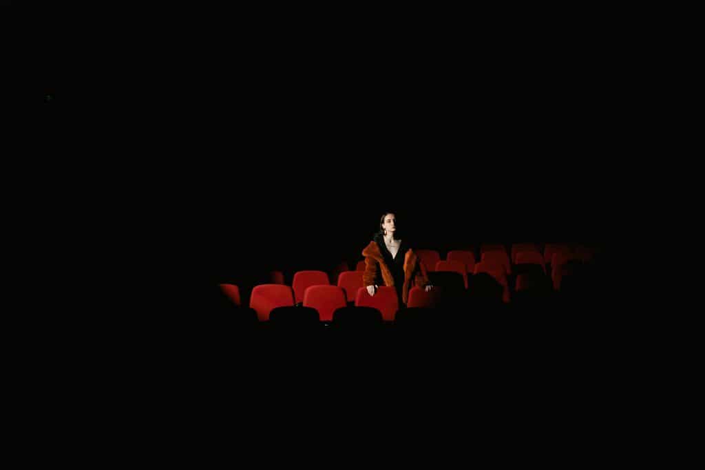 A woman in a fur coat sits alone in a dimly lit theater, surrounded by empty red seats.