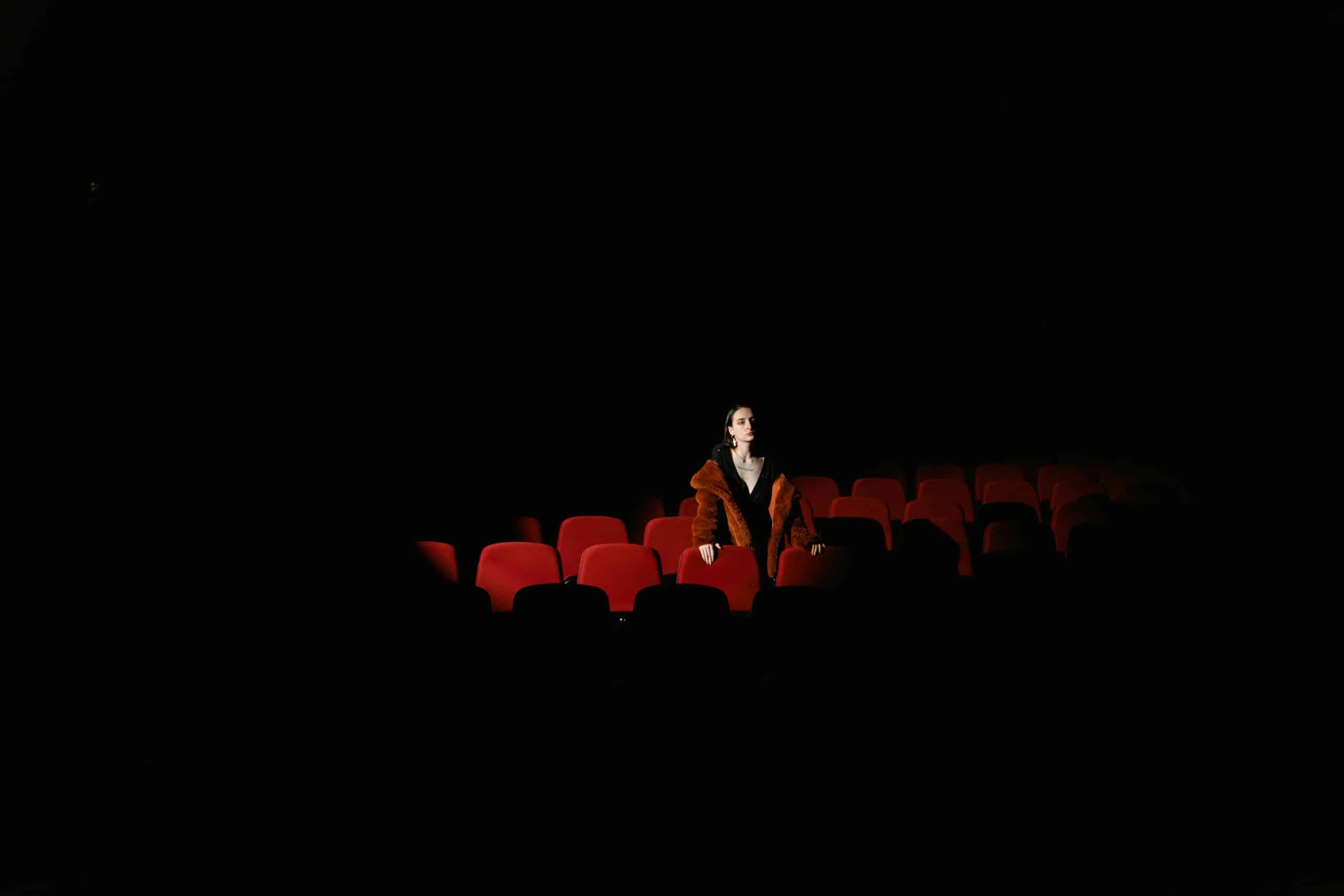 A woman in a fur coat sits alone in a dimly lit theater, surrounded by empty red seats.