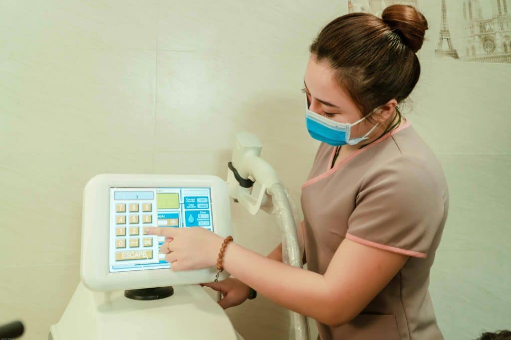 A healthcare worker in a mask operates a medical touchscreen device in a clinic setting.